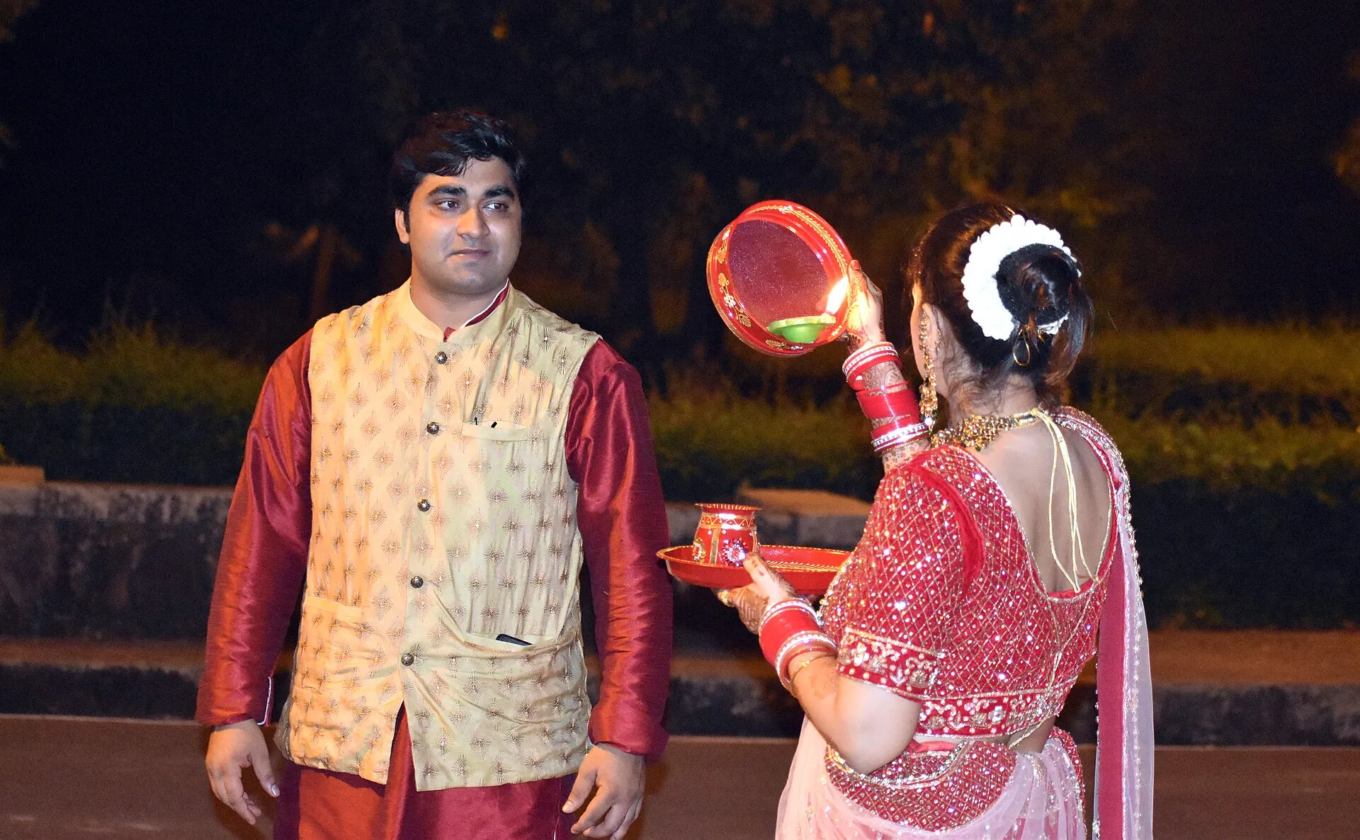 Woman in red bridal dress holding a sieve and puja thali during Karva Chauth at night