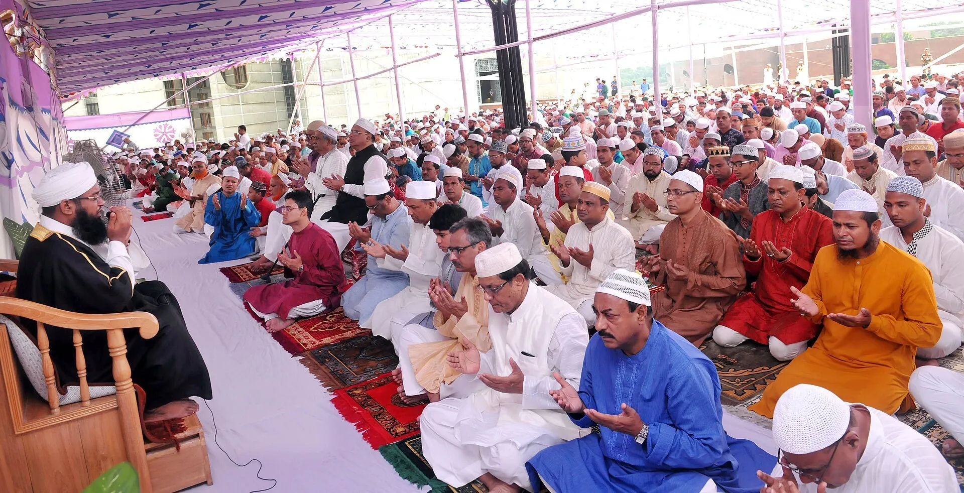 A large outdoor congregation in festive dress with hands raised in prayer at Eid al-Fitr