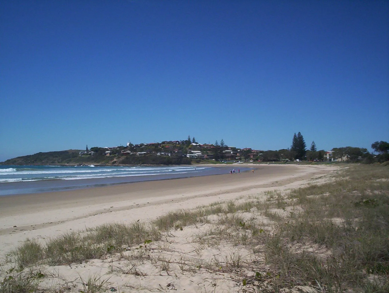 Woolgoolga main beach with golden sand and surf, the town and headland in the distance under a clear blue sky