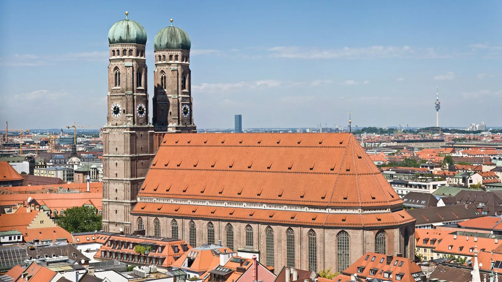 The Frauenkirche in Munich, its twin onion domed towers and red roof above the old town rooftops under a blue sky